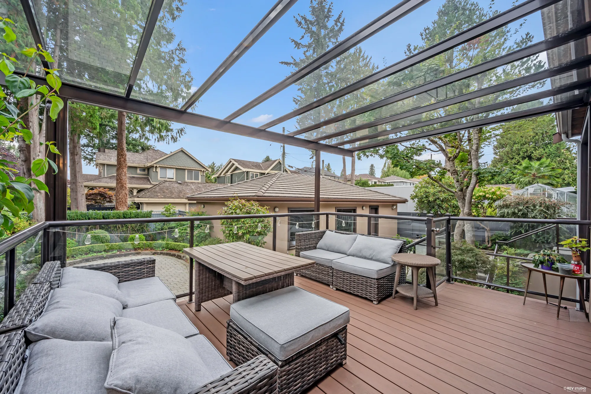 Under the glass pergola: a wicker sofa set on the composite deck, the garden and the neighbours' roofs beyond the rail