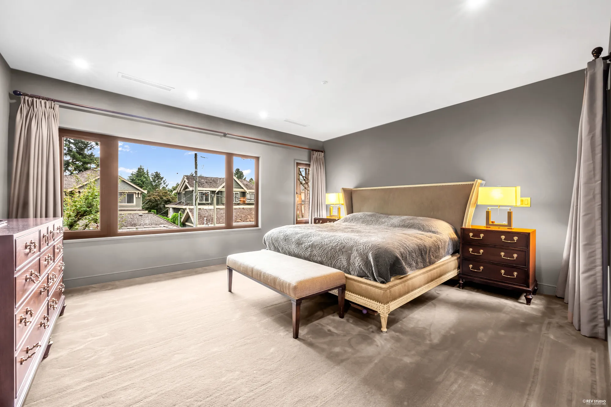 The primary bedroom: the bed facing a wide window, taupe walls, a bench at its foot