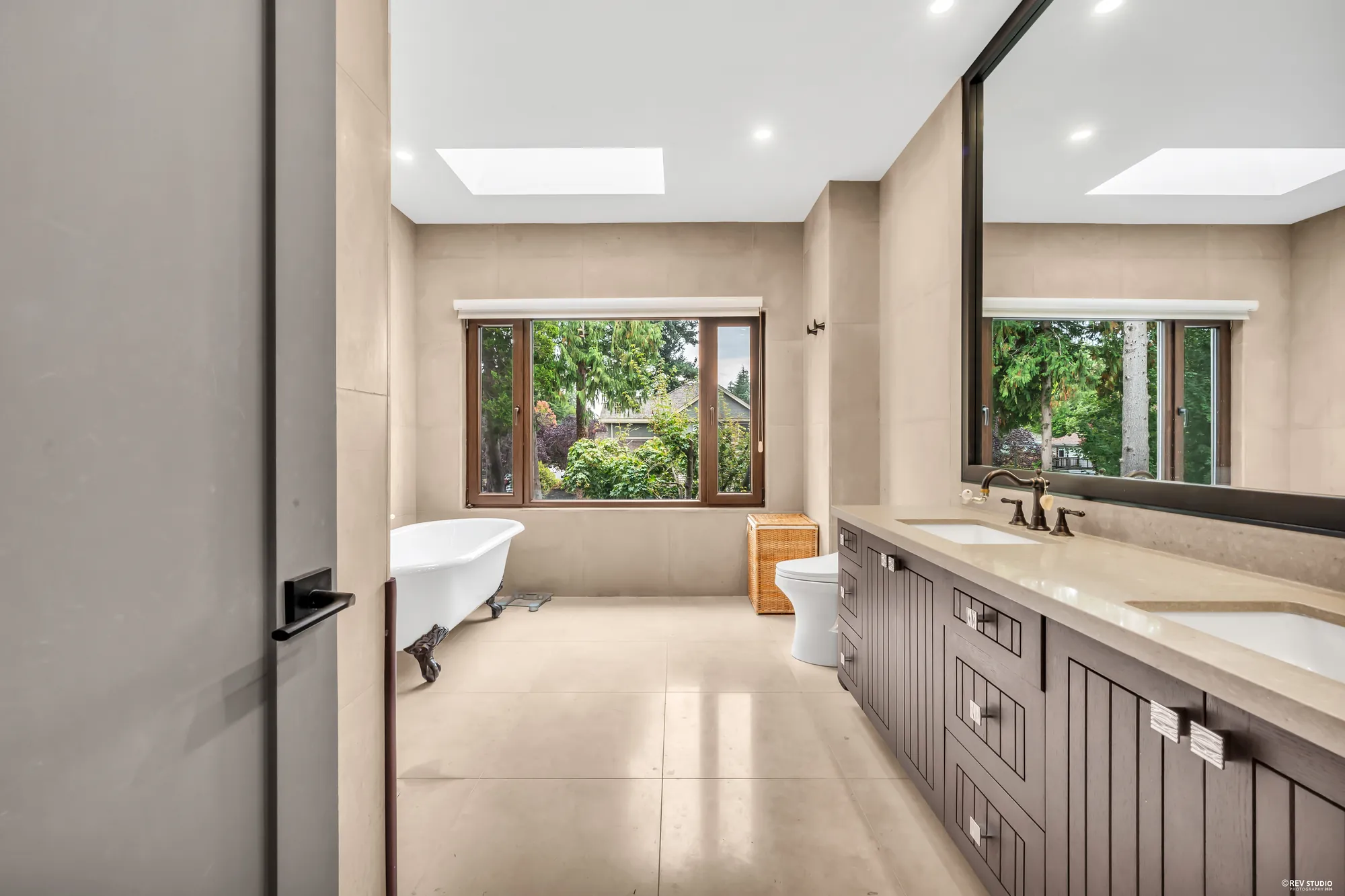 The primary bathroom: a freestanding tub in the window under a skylight, a double vanity in dark wood with a stone top, a large mirror
