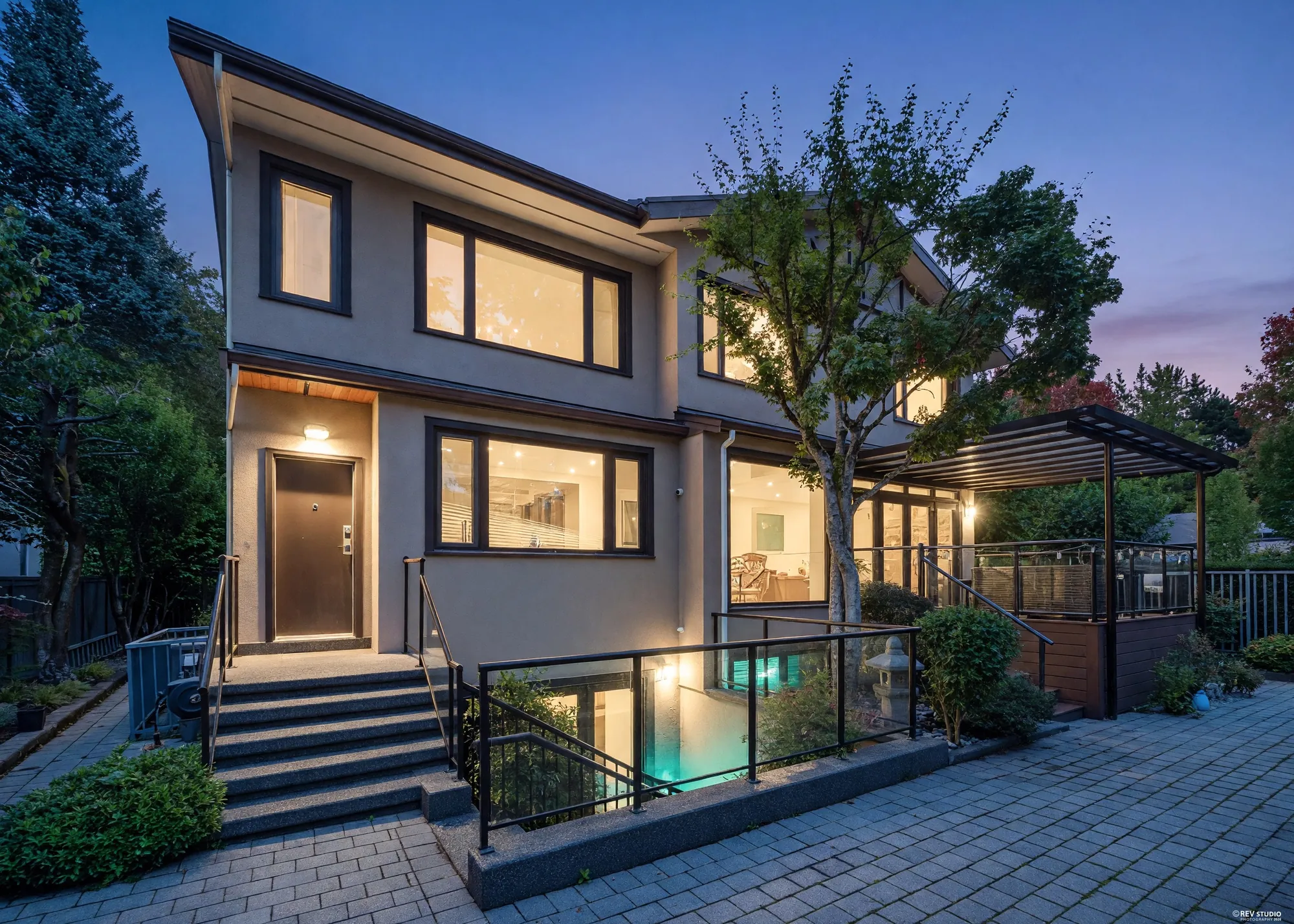 The back of the house at dusk: stucco and brick, every window lit, the pergola over the deck at the right and the stair down to the lower patio