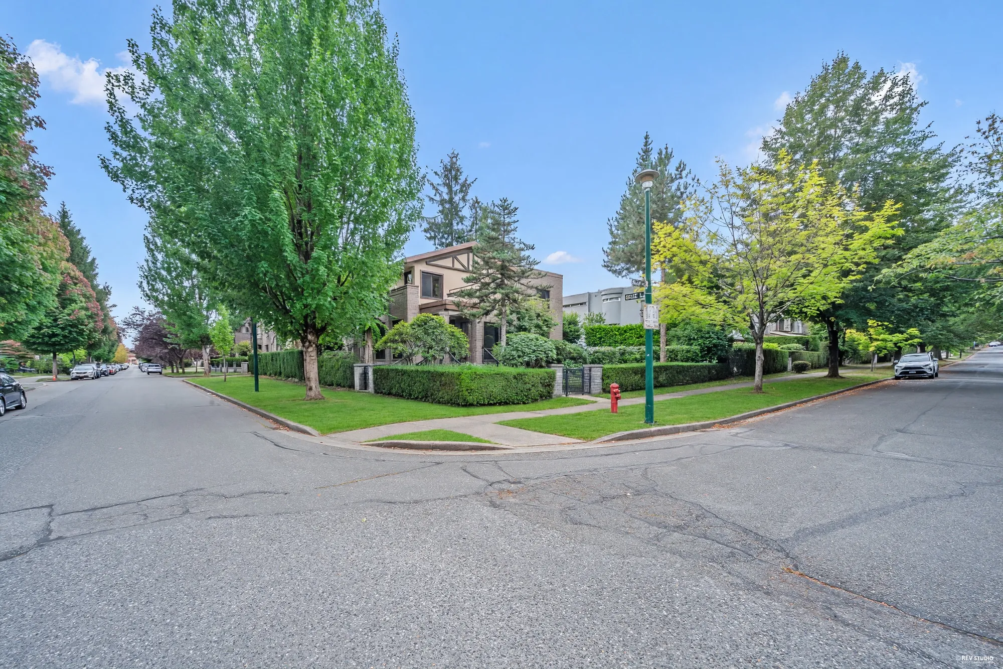 The house from across the intersection under the street trees, the hedge running along both streets